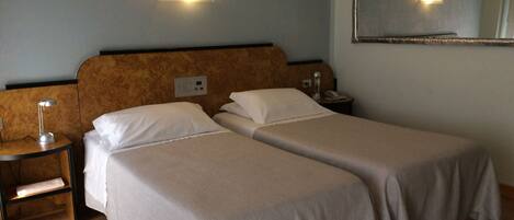 A hotel room with two single beds, a nightstand with a lamp, a bedside table with a phone, and a wall-mounted light fixture.