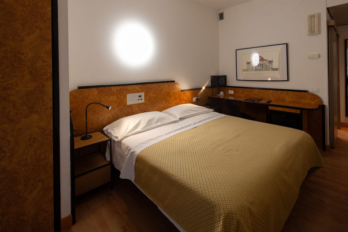 Una habitación de hotel con cama, mesita de noche, lámpara y un cuadro enmarcado en la pared.