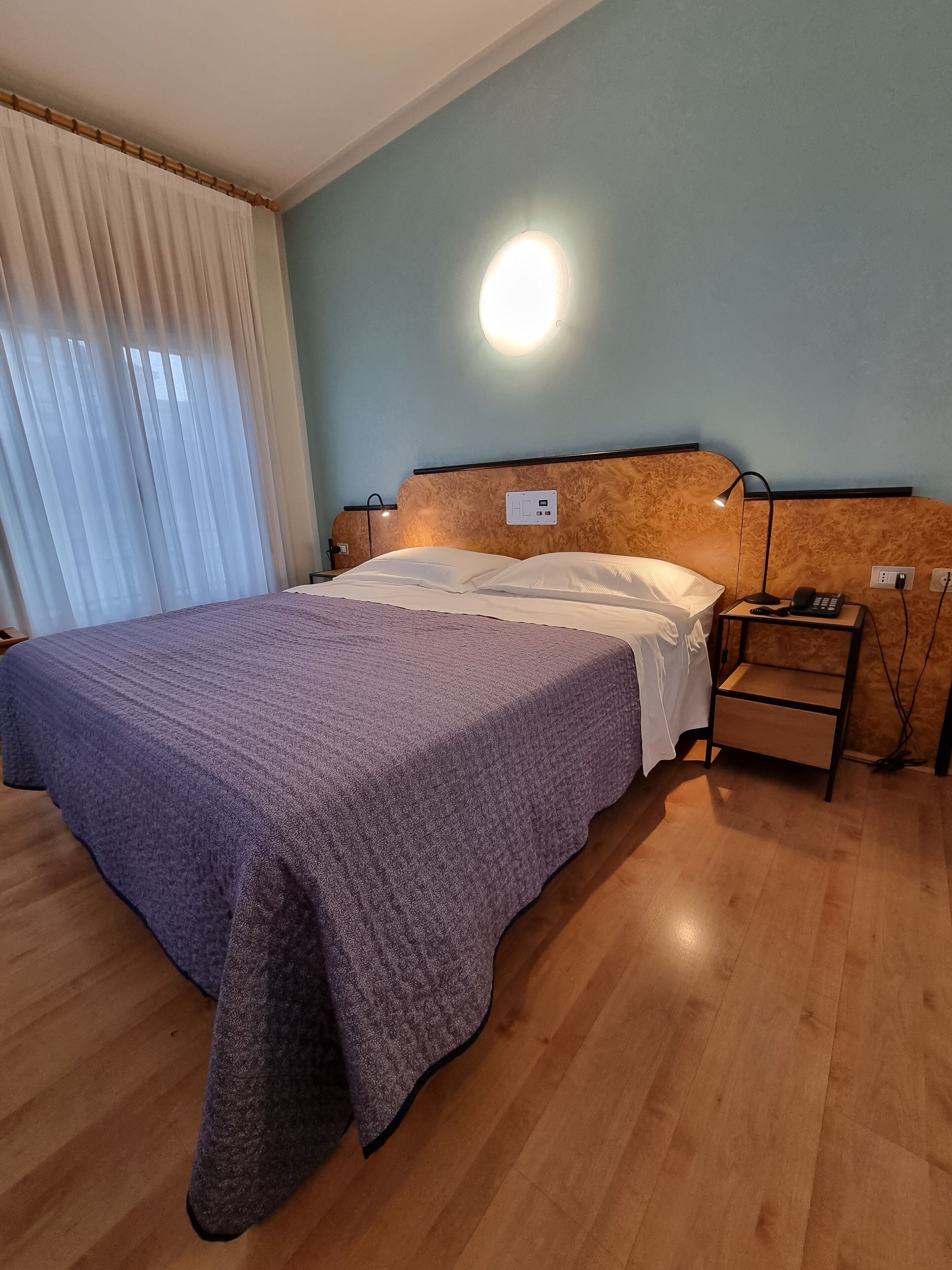 A neatly made bed with a purple blanket, a bedside table with a lamp, and a wall-mounted light fixture.
