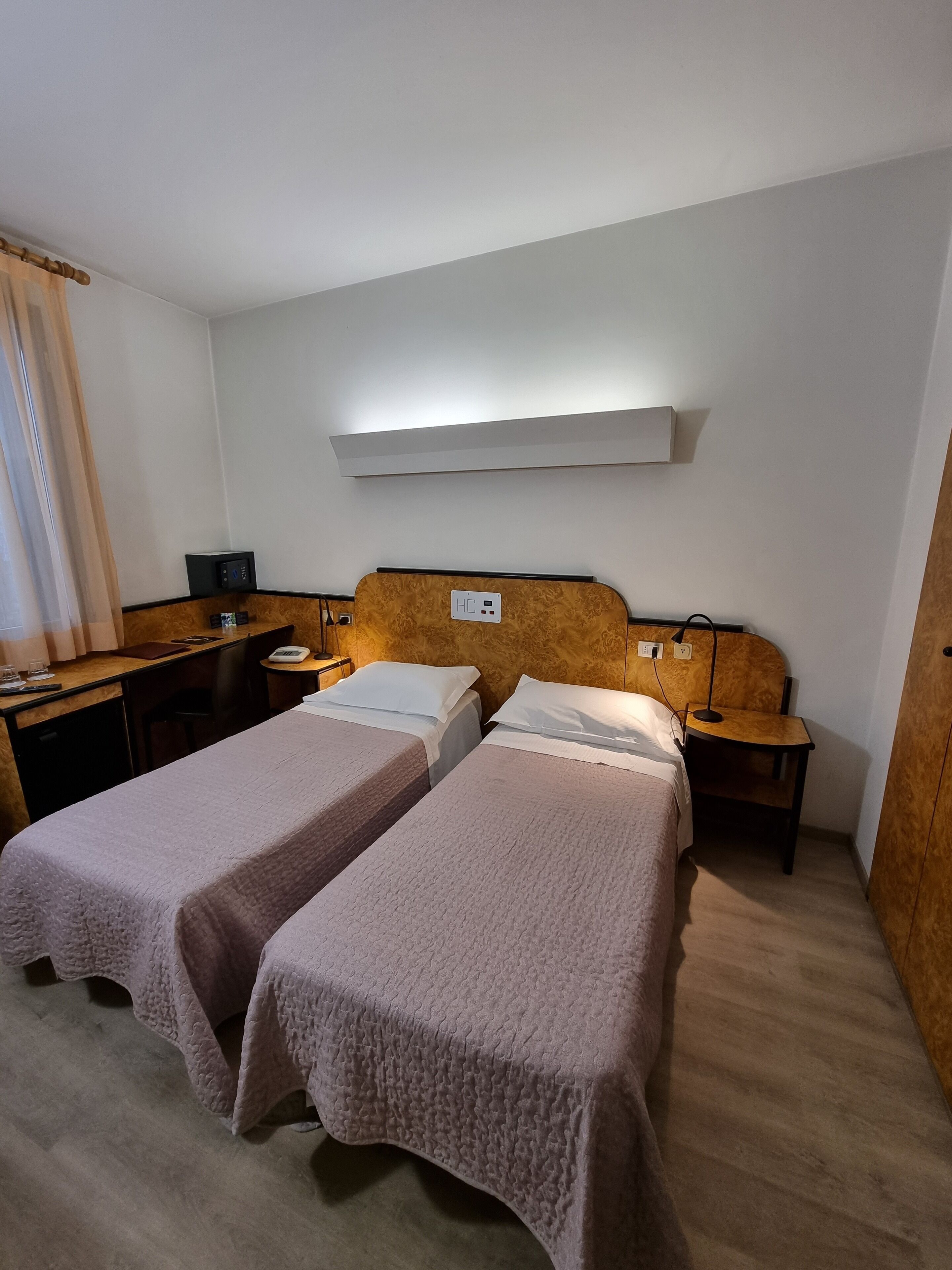 A hotel room with two single beds, a desk, a chair, a small table, and a nightstand with a lamp.