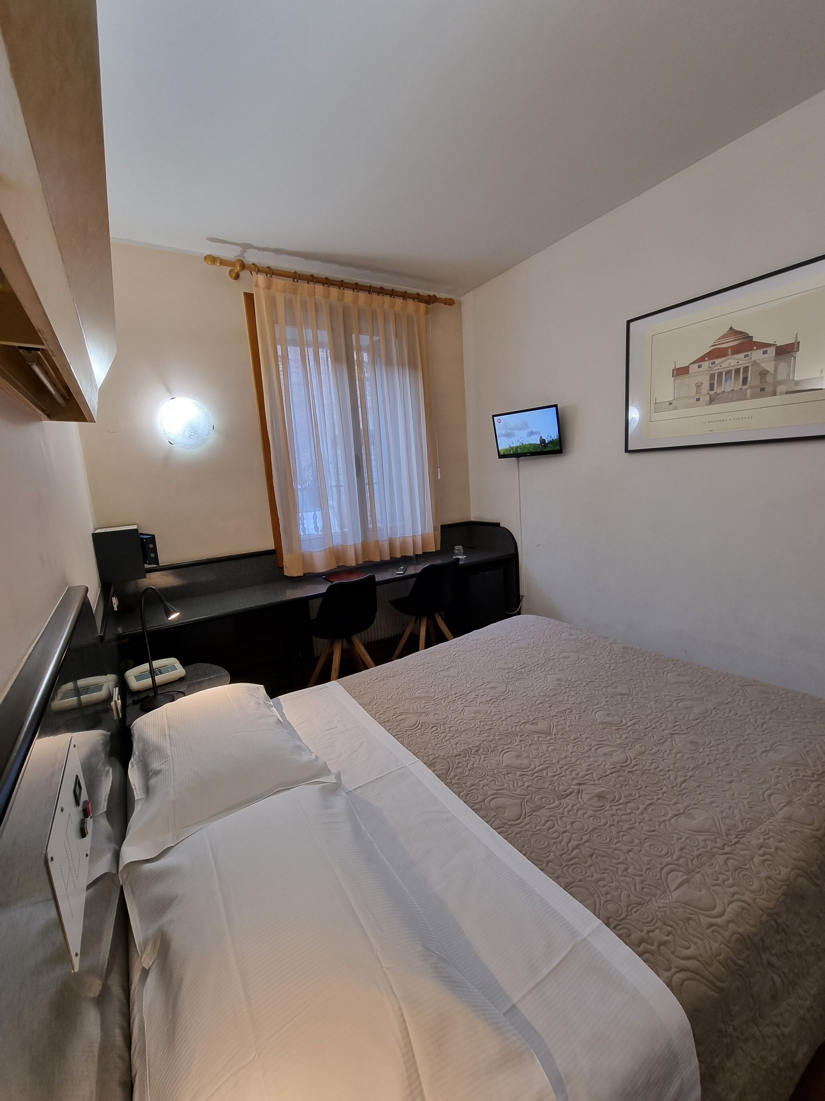 Habitación de hotel con una cama, un escritorio con silla, un televisor y un cuadro en la pared.