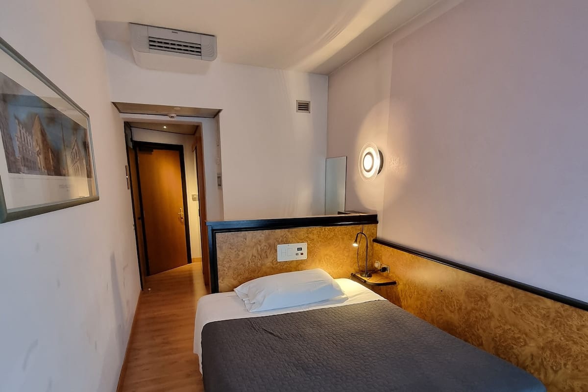 Habitación de hotel con cama, mesita de noche, luz empotrada en la pared y puerta que da a otra habitación.