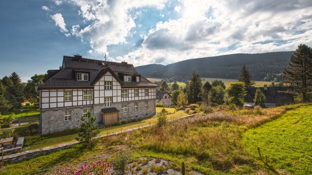 A traditional half-timbered house with a stone foundation, surrounded by a lush green landscape and a mountainous backdrop. - Theresian Apartment (Ostruzna)