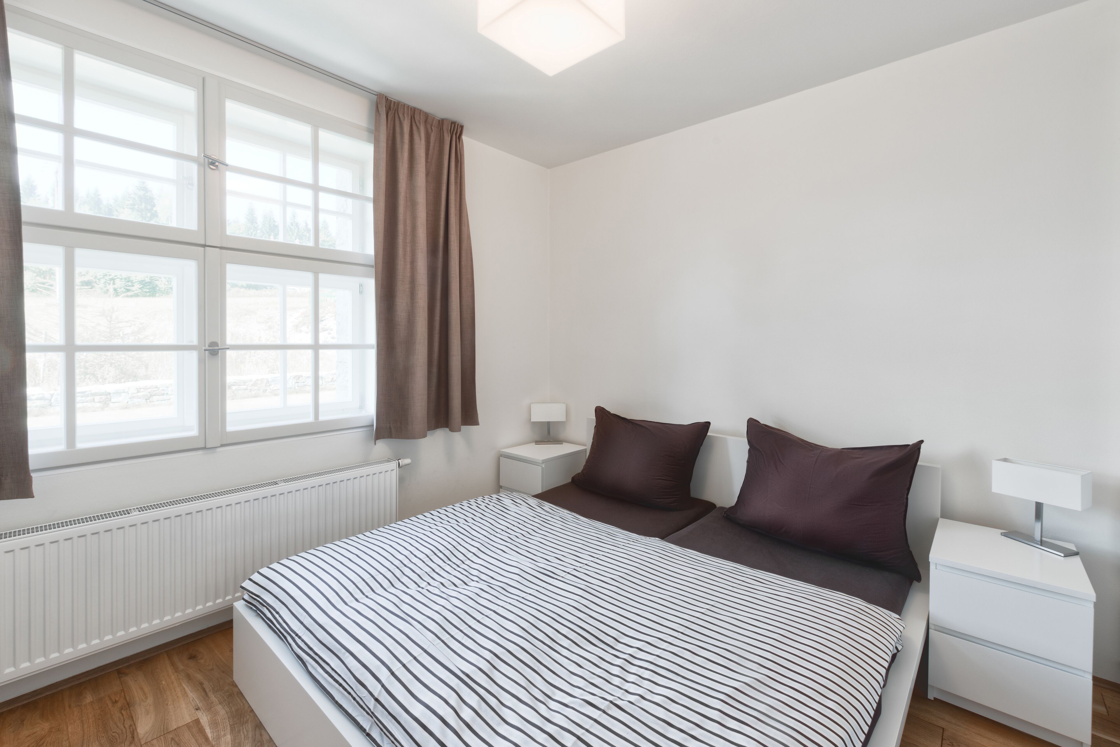 A bedroom with a bed, a nightstand, a lamp, a window with curtains, and a radiator.
