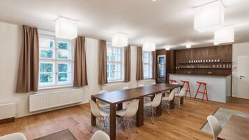 A spacious room with a long wooden table, white chairs, and a kitchen area with stools.