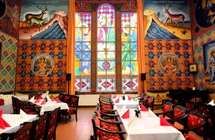 Un comedor con mesas y sillas, un amplio ventanal con vitrales y arte religioso en las paredes.