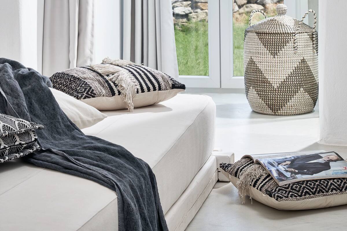 Una sala de estar moderna con un sofá blanco, un plaid estampado y una revista sobre un cojín. Una cesta tejida se encuentra cerca de un ventanal que da a un balcón con vistas a un paisaje rocoso.