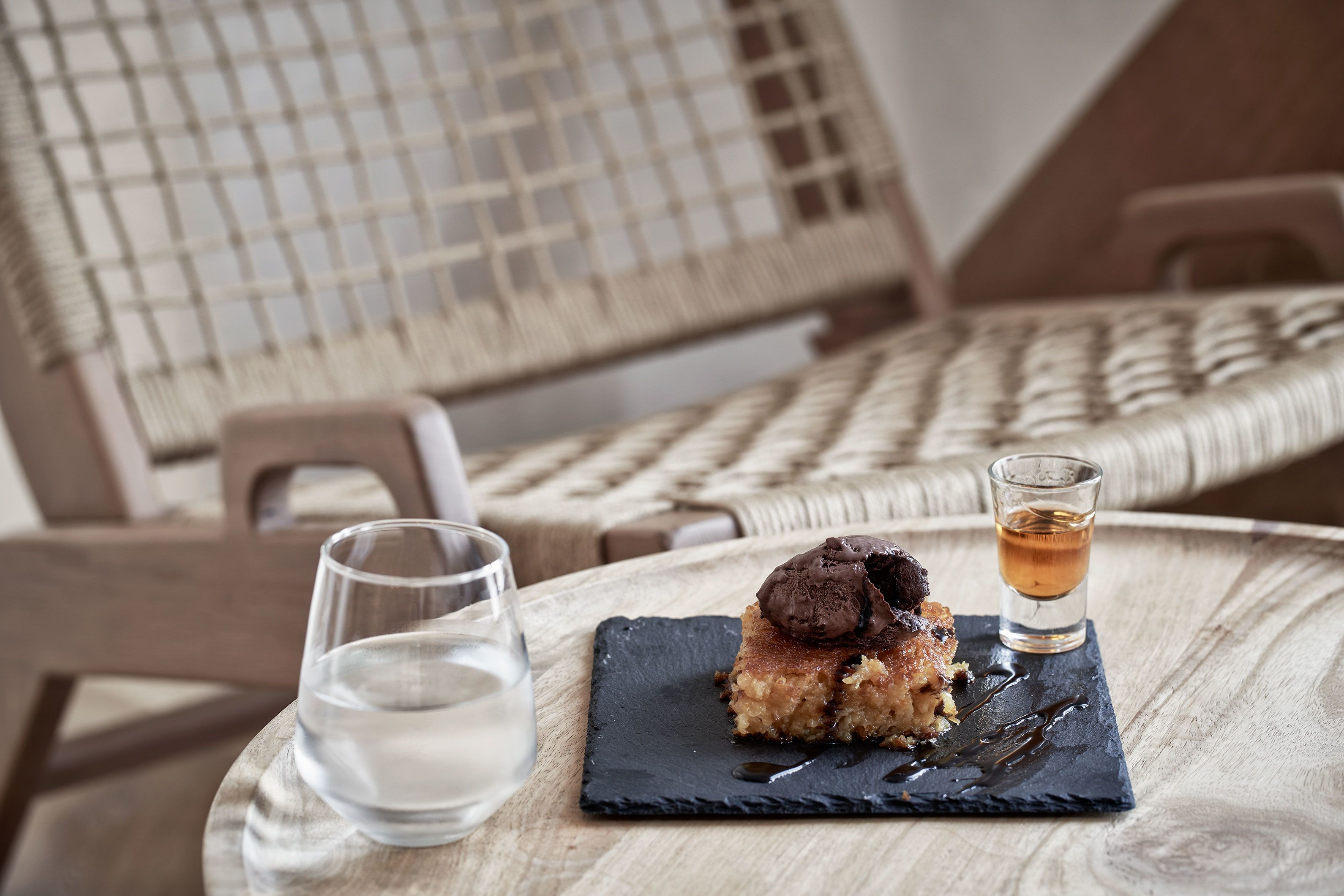 A dessert on a slate plate with a shot of whiskey on the side, a glass of water, and wicker chairs in the background.