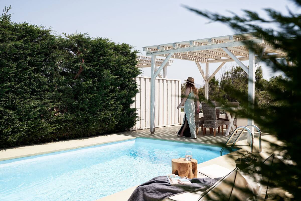 Un área de piscina con una pérgola, sillas de descanso y un taburete de madera con un vaso.
