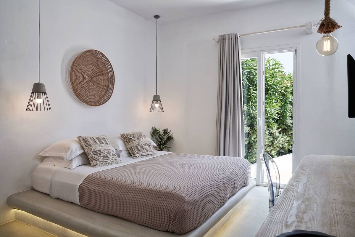 Un dormitorio moderno con cama, lámparas de noche, un adorno circular tejido en la pared y vistas a la vegetación a través de una ventana.