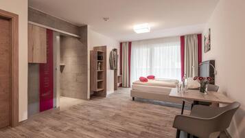 Una habitación de hotel moderna con suelo de madera, una cama con almohadas rojas, un escritorio con una silla y un televisor en la pared.