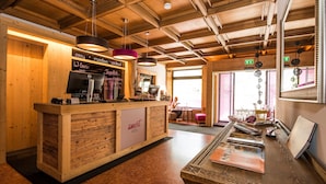 A reception desk with a wooden finish, a digital screen, and a sign reading "smart hotel". There are hanging pendant lights, a window with a view, and a door with an exit sign. - smartHOTEL (Dorfgastein)