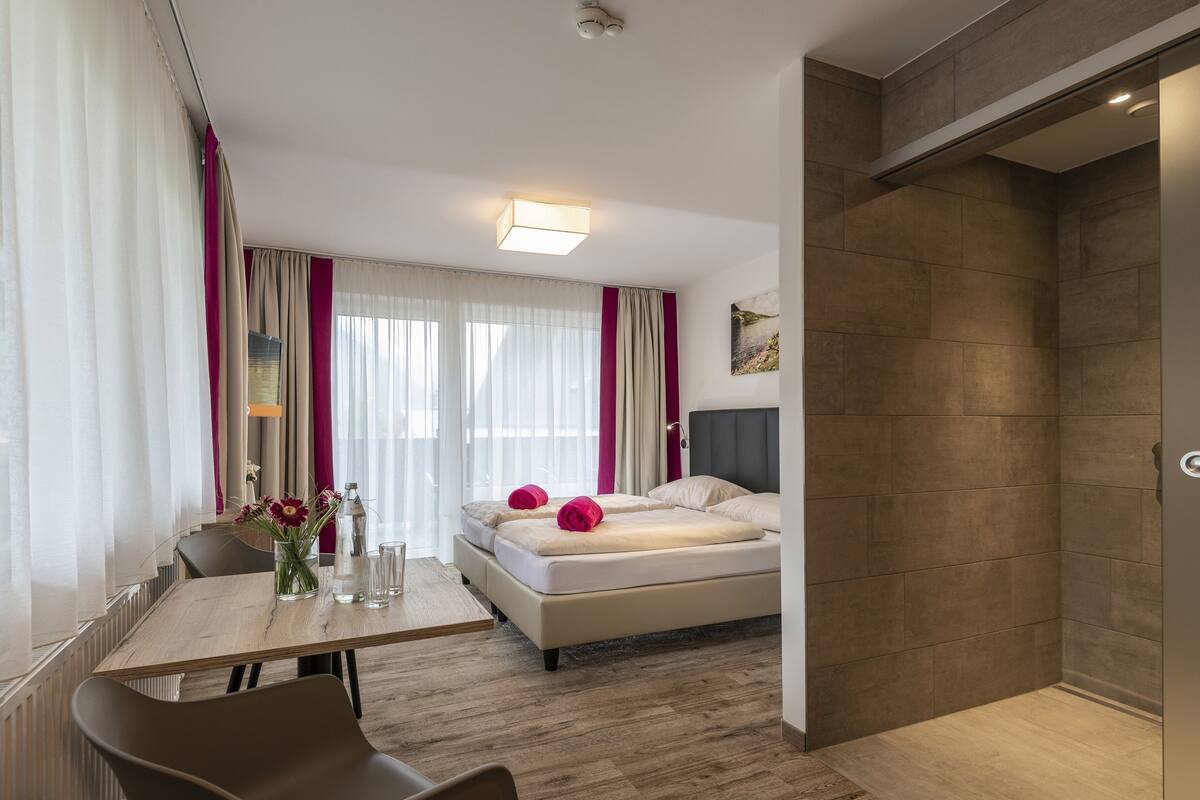 A modern hotel room with a bed, a desk with a chair, a shower area, and a window with curtains.