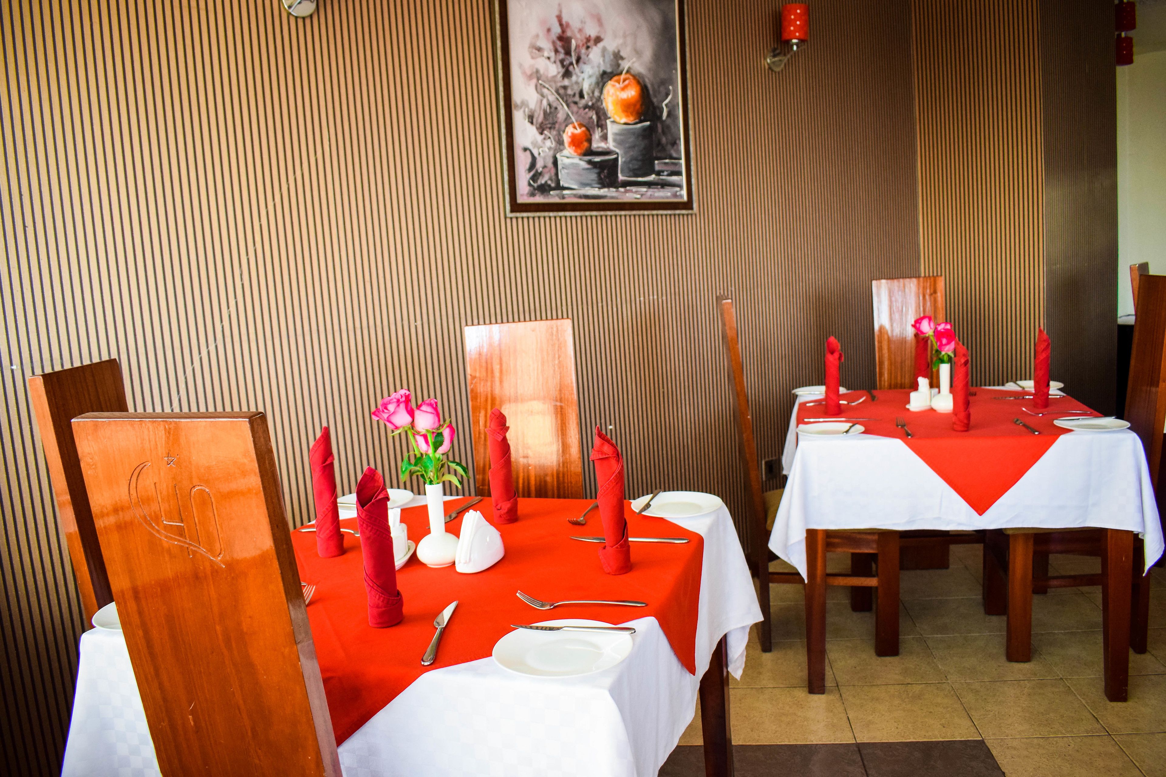 A dining area with tables set for two, featuring red napkins, white tablecloths, and a framed painting on the wall.