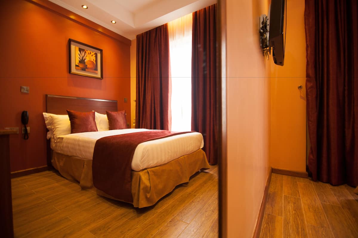A hotel room with a large bed, wooden flooring, a painting on the wall, and red curtains.
