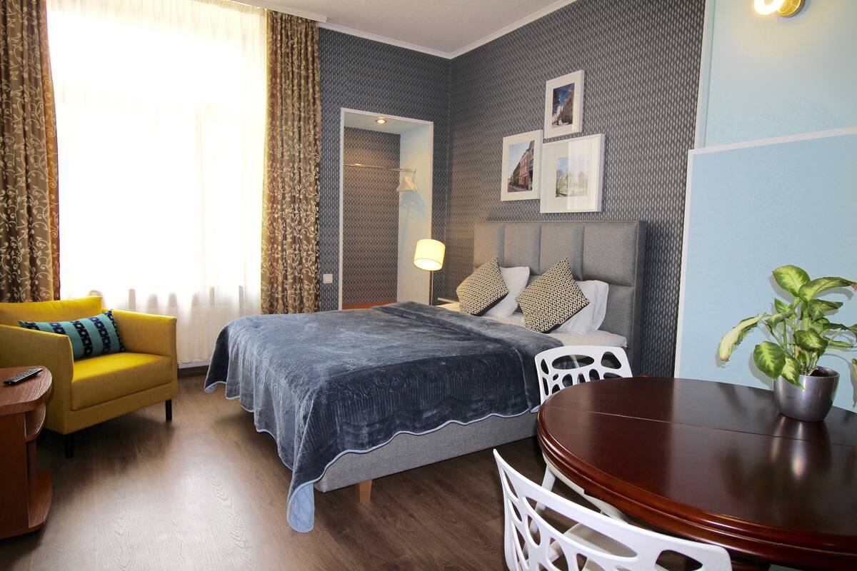 A hotel room with a bed, a yellow armchair, a wooden table, and a potted plant.