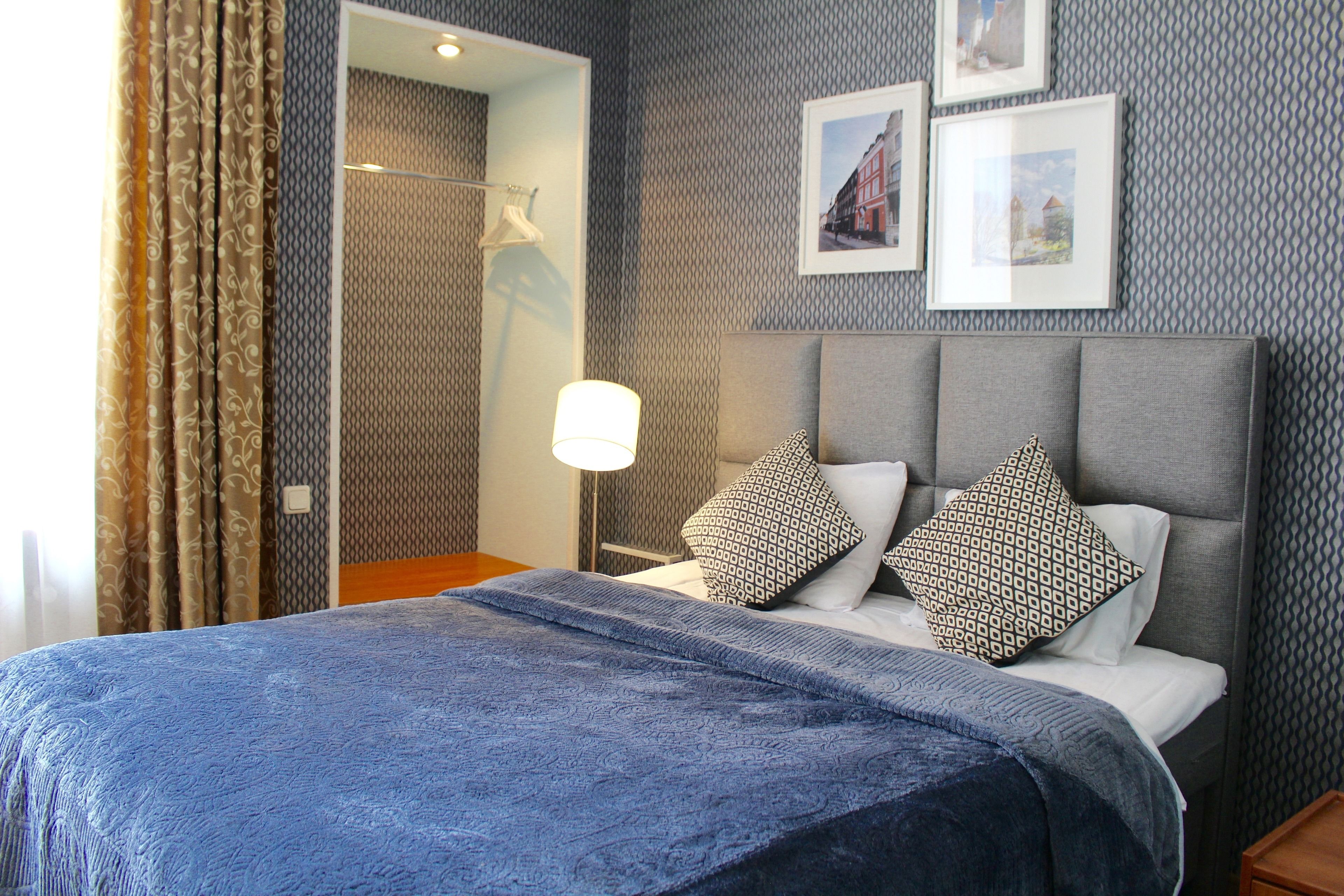 A hotel room with a bed, a nightstand, a lamp, and a wall with patterned wallpaper and framed pictures.