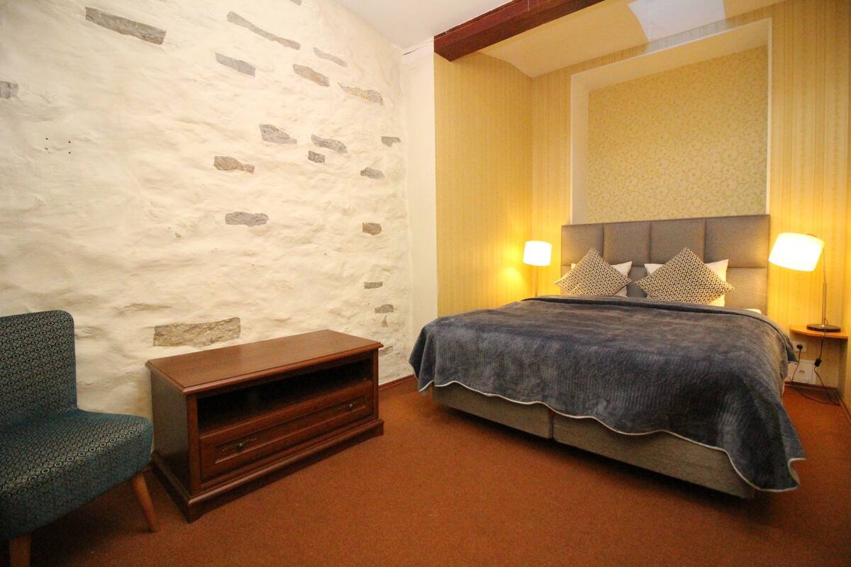 A bedroom with a stone wall, a bed with a grey bedspread, two bedside lamps, a wooden nightstand, and a patterned chair.