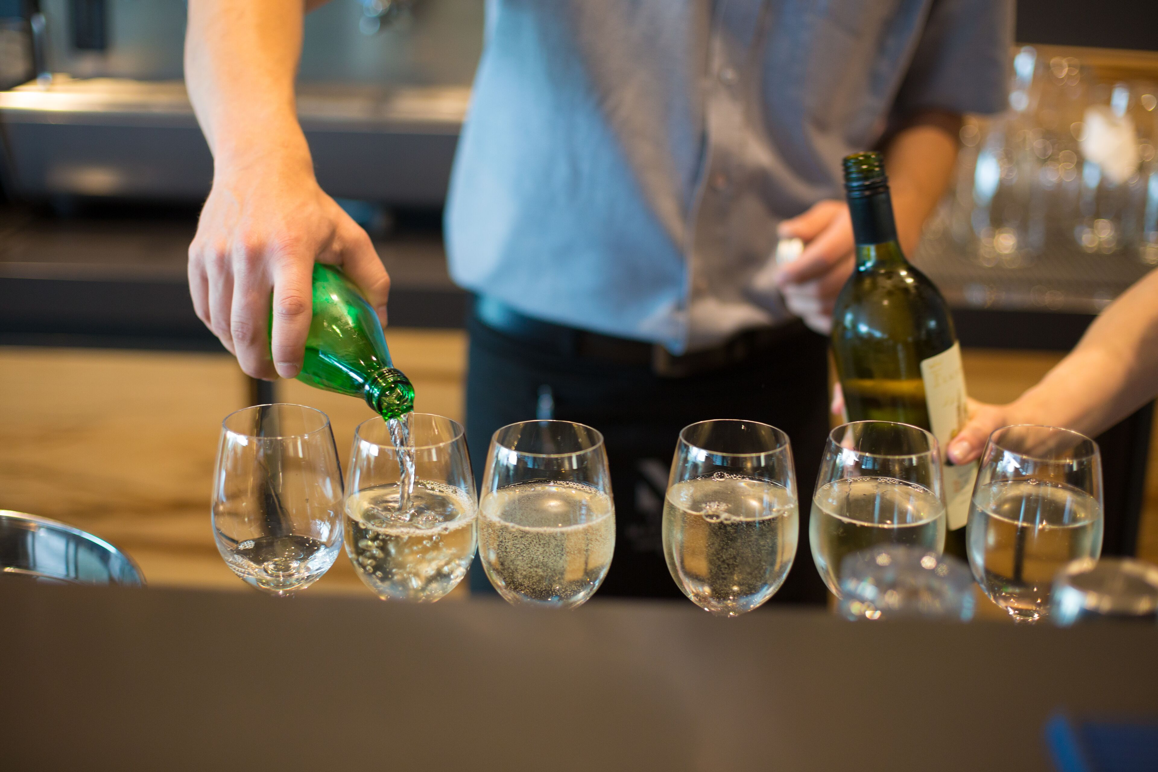 Eine Person gießt Flüssigkeit aus einer grünen Flasche in mehrere Gläser hinter einer Bar.