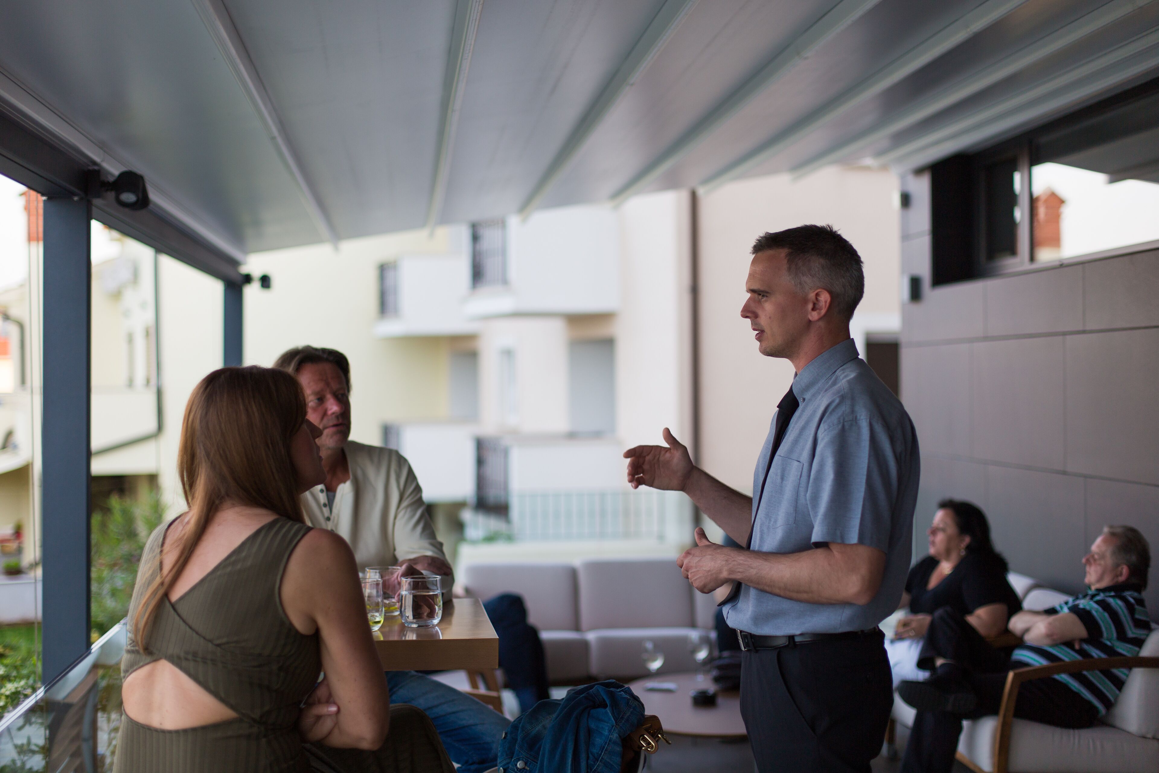 A modern outdoor seating area with a covered patio, a table with a glass of water, and a person standing and talking to another person.