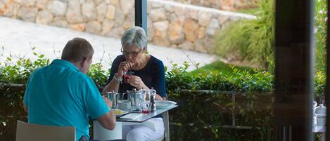 Dos personas sentadas en una mesa al aire libre, con un muro de piedra y follaje de fondo.