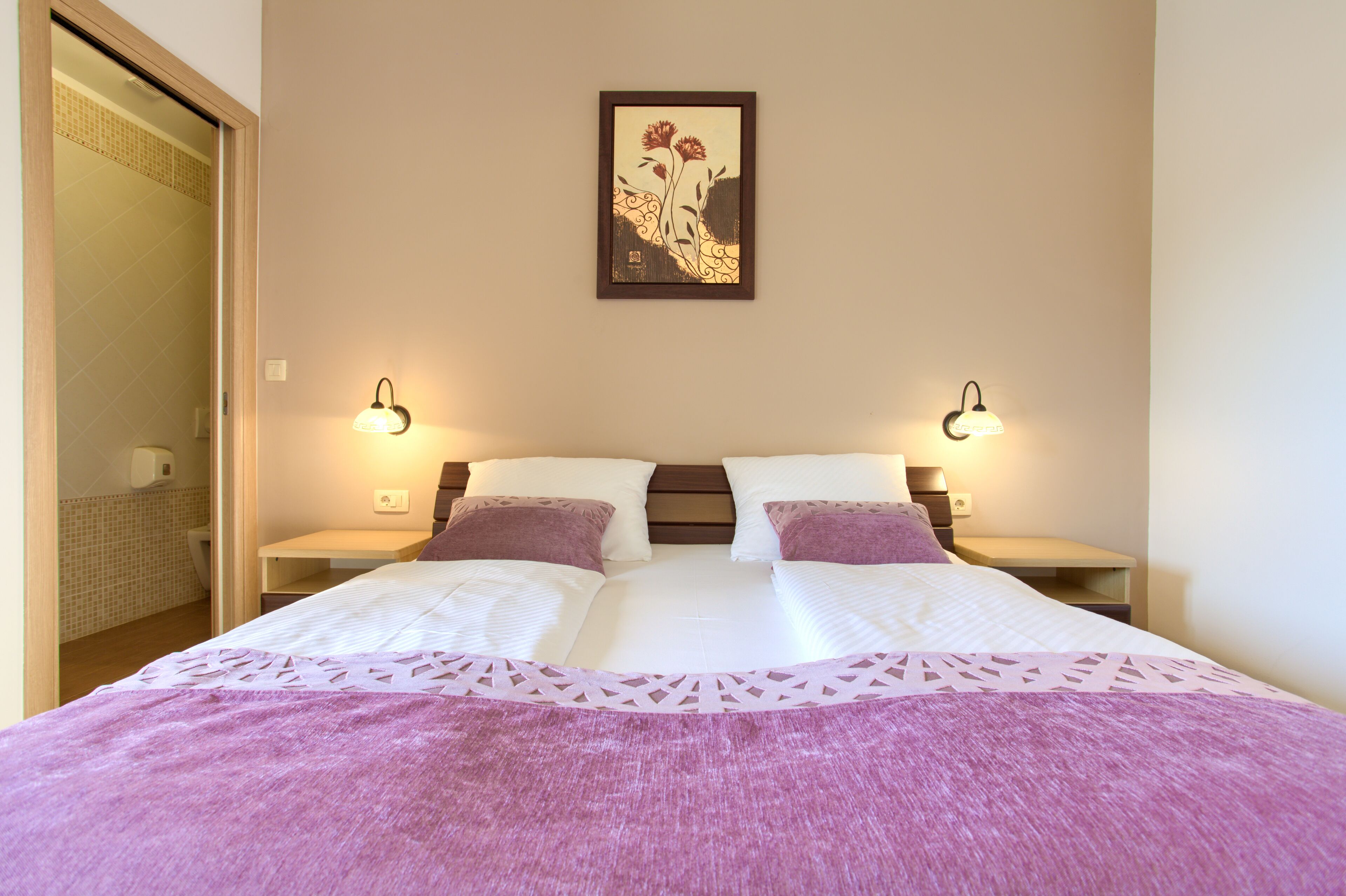 A hotel room with a bed, two nightstands, a framed artwork, and a bathroom visible through an open door.