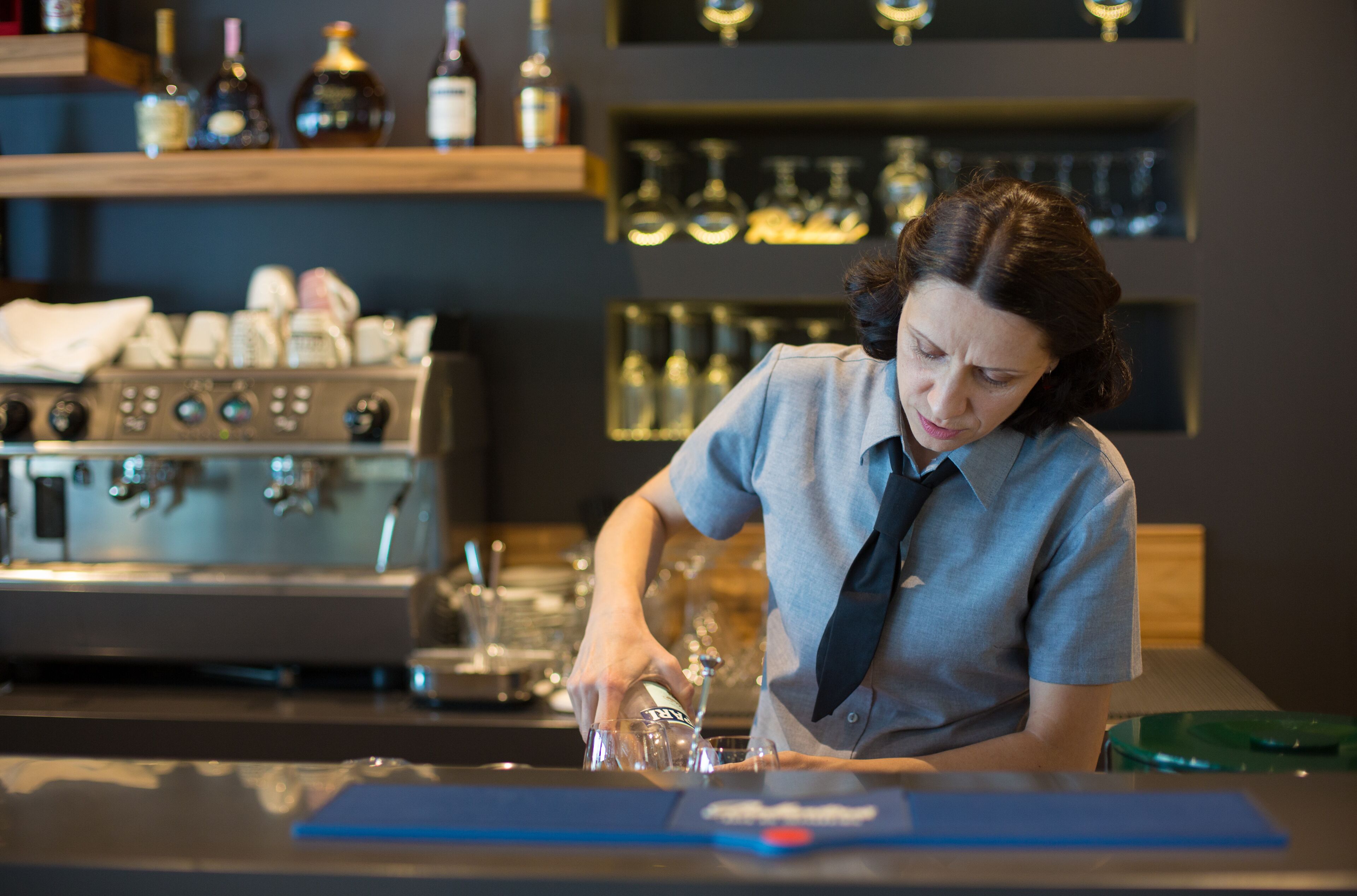 Un barista preparando una bebida en la barra, con una máquina de café y estantes llenos de botellas al fondo.