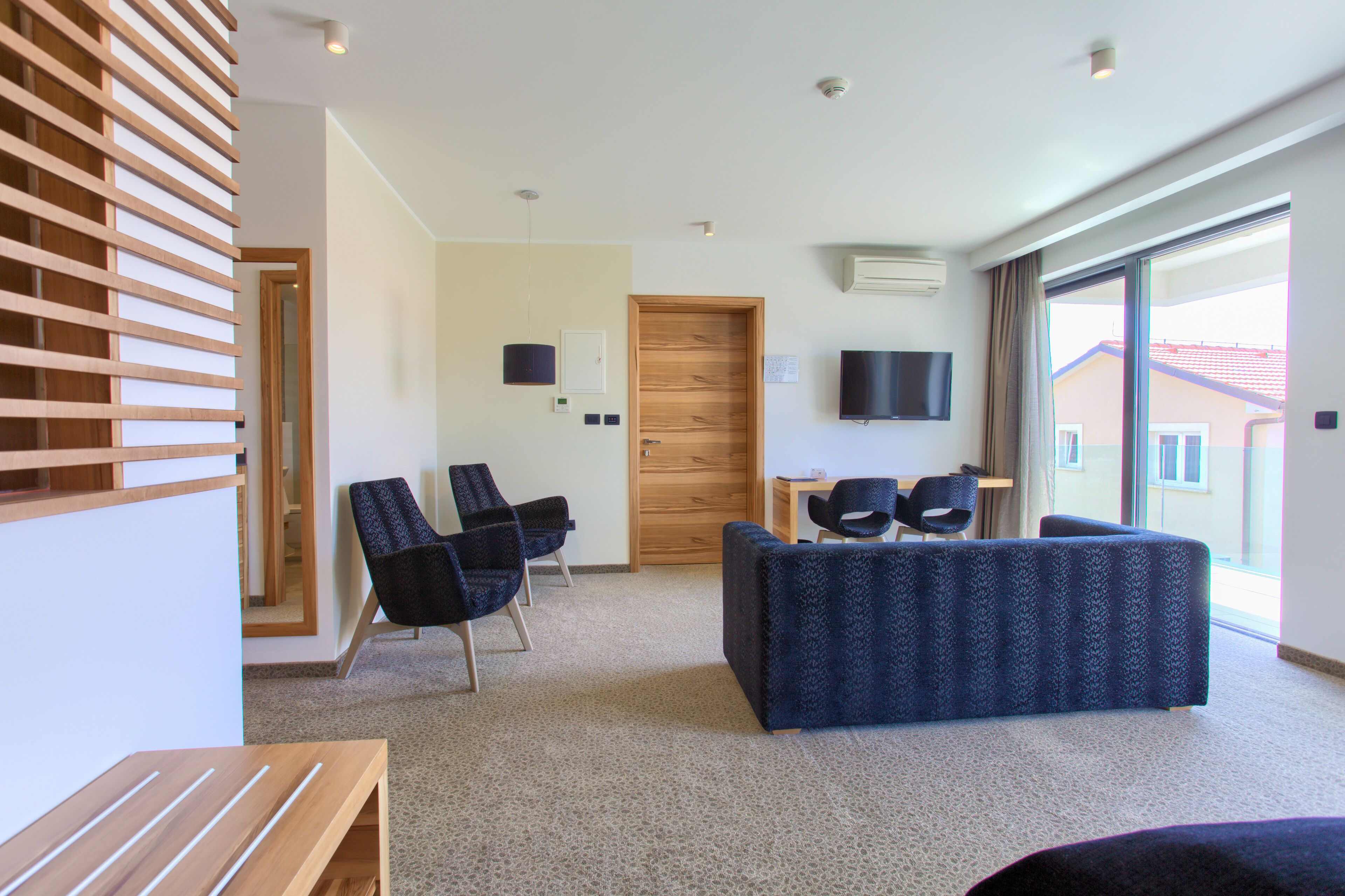 Een moderne hotelkamer met een wand van houten latten, een groot raam met uitzicht, een flatscreen televisie en een zithoek met stoelen en een bank.