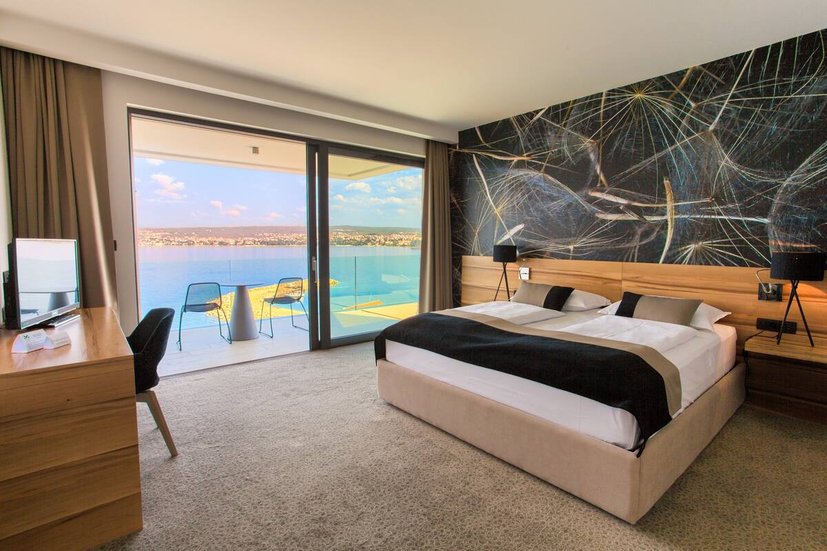 Ein Hotelzimmer mit einem großen Bett, einem Schreibtisch mit Computer, einem Stuhl und Meerblick durch eine Glastür.