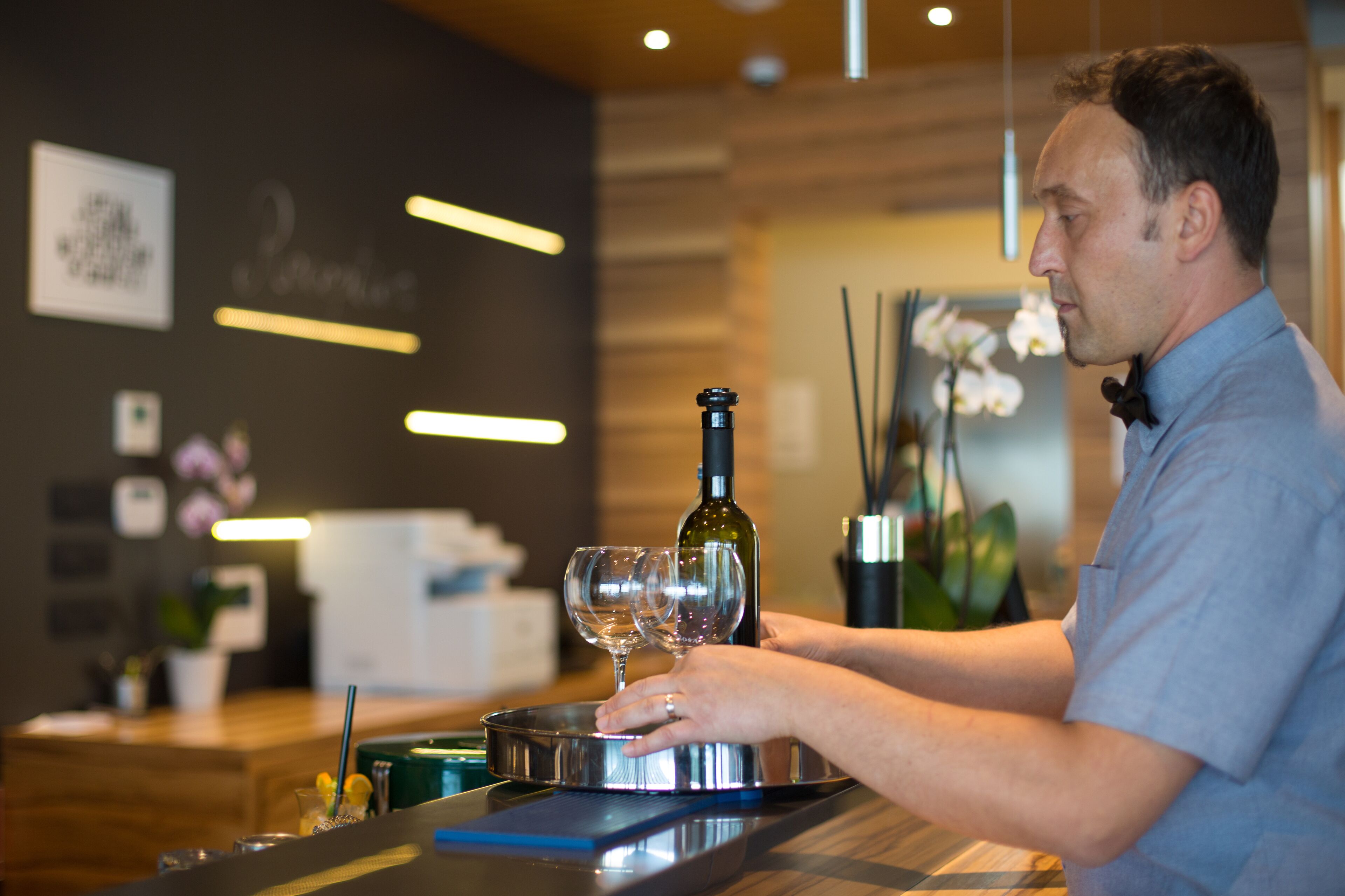 A person pouring wine into a glass at a bar with a modern interior, including a wooden wall and a framed picture.