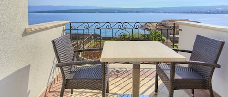 A balcony with a table and two wicker chairs, overlooking a body of water and distant mountains.