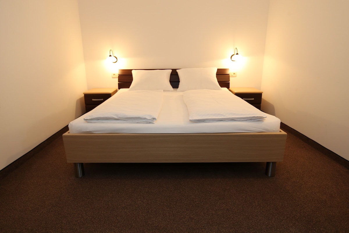 Ein ordentlich bezogenes Bett mit weißen Laken, zwei Nachttische mit Lampen und ein Teppichboden in Braun.