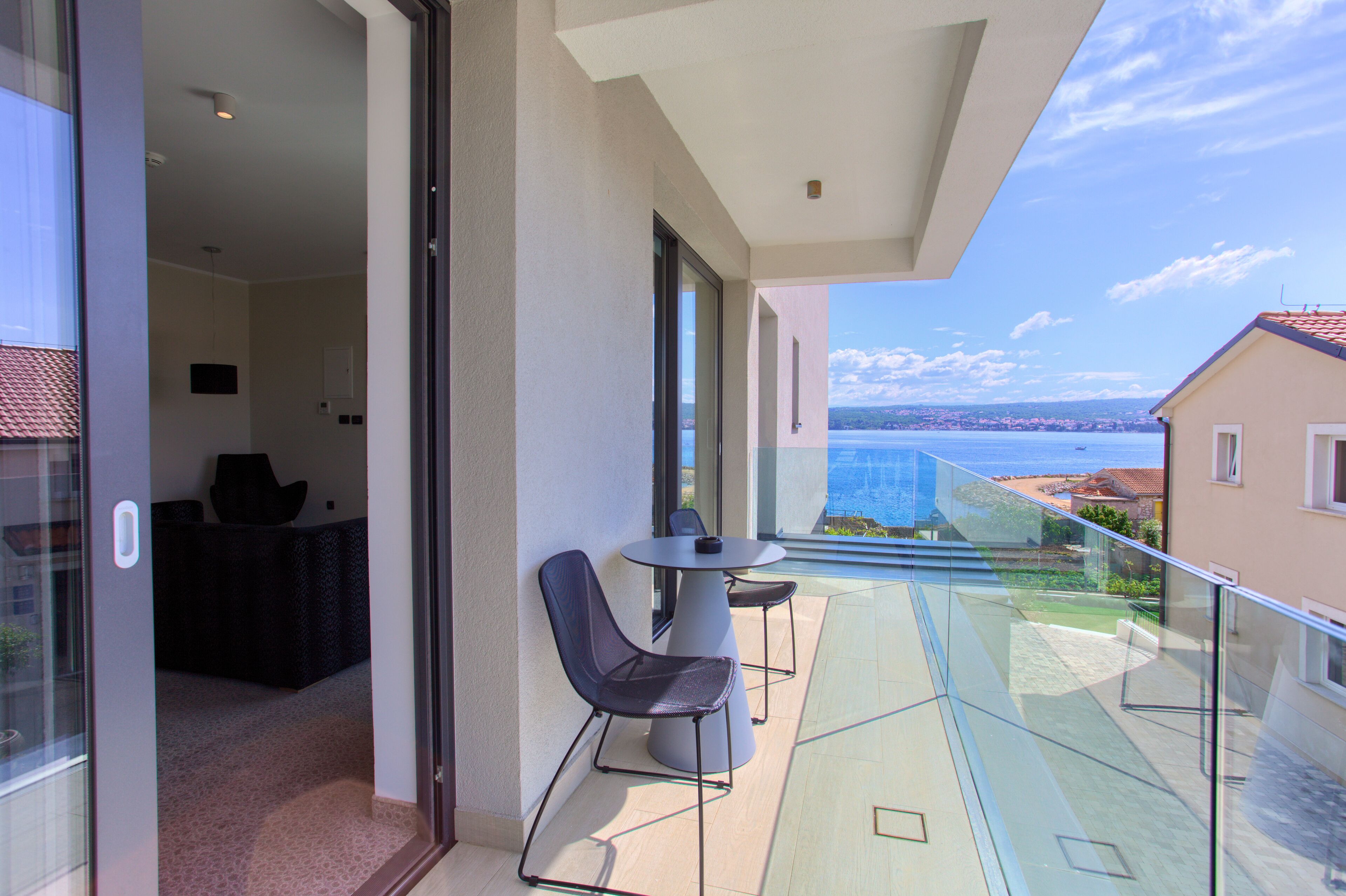 A balcony with a glass table and chairs, a view of the sea, and a building with a red-tiled roof.
