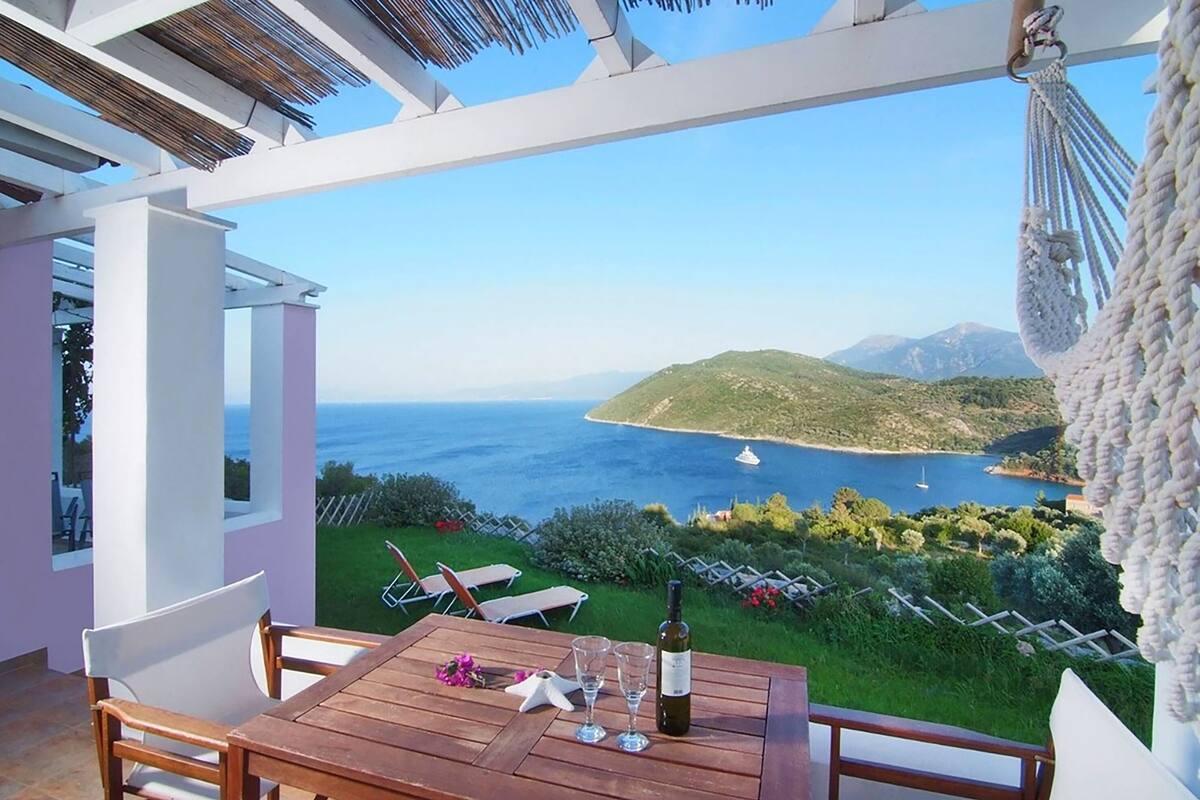 Une table en bois, avec une bouteille et des verres, donnant sur un paysage côtier offrant une vue dégagée sur la mer et les collines.