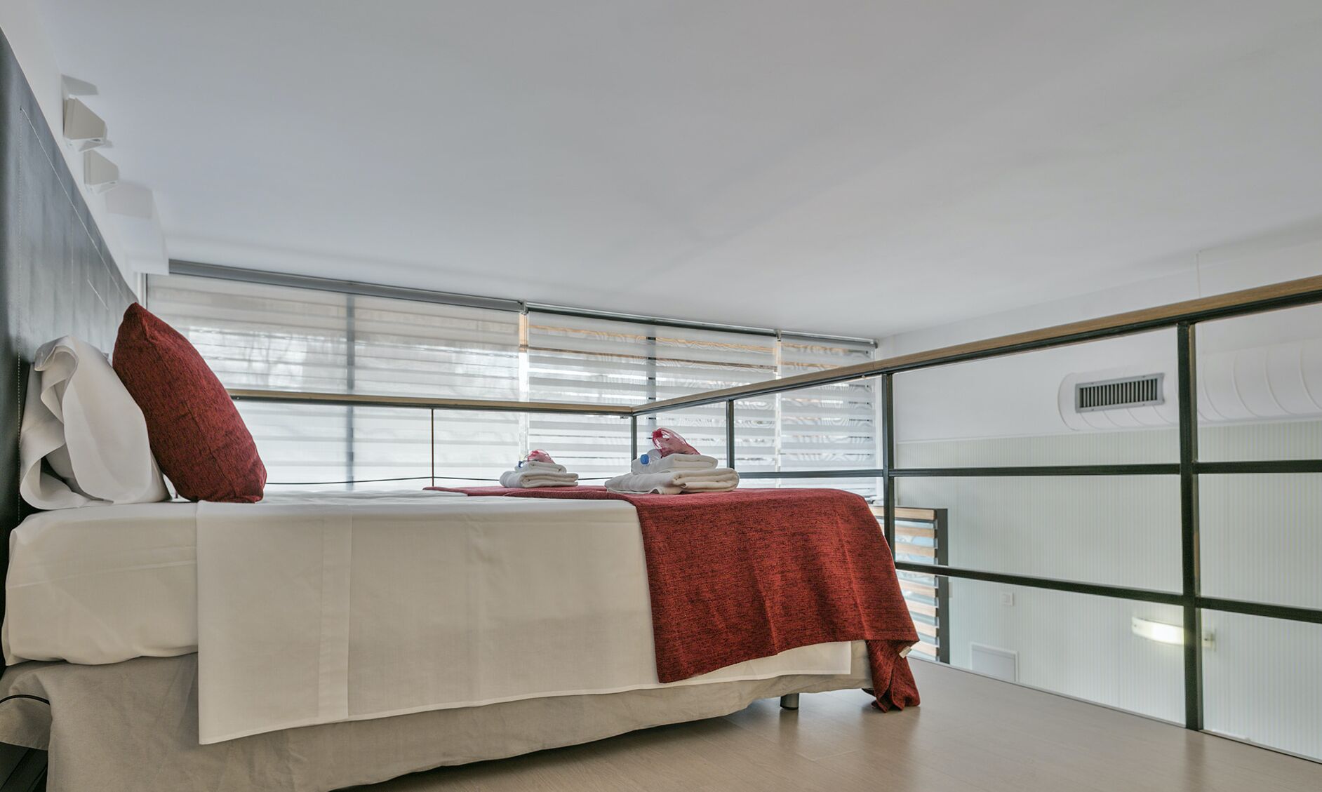 A modern bedroom with a bed, a red blanket, and a view of the outside through large windows.