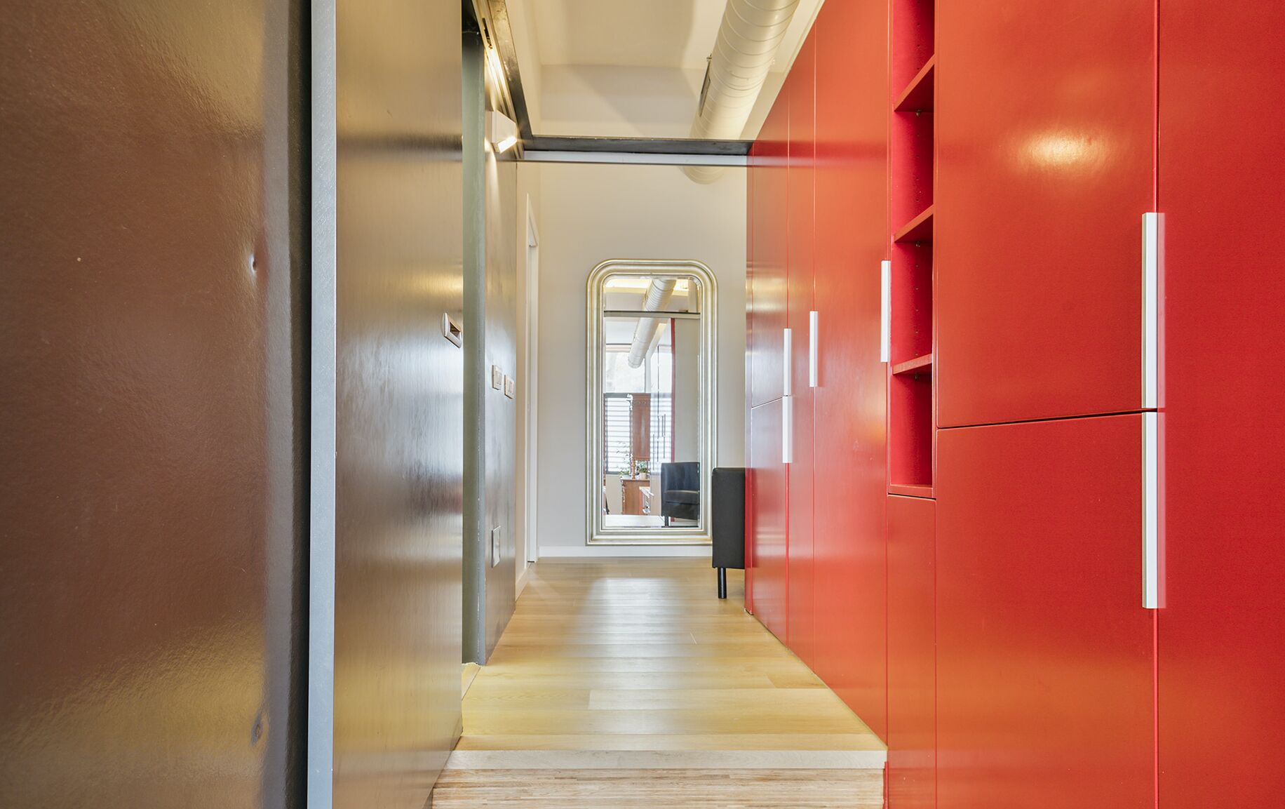 A modern hallway with a wooden floor, a stainless steel door, and red cabinets with a mirror.