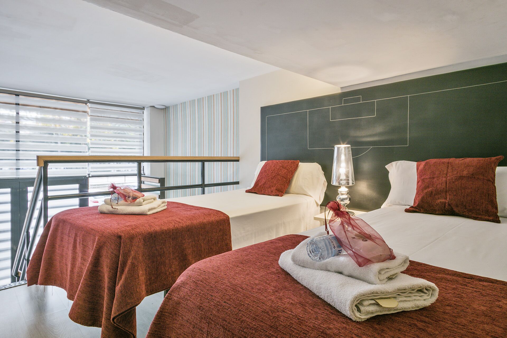 Habitación de hotel con dos camas, cada una con una toalla roja y una botella de agua. Se observa una mesita de noche con una lámpara, y una ventana con persianas.