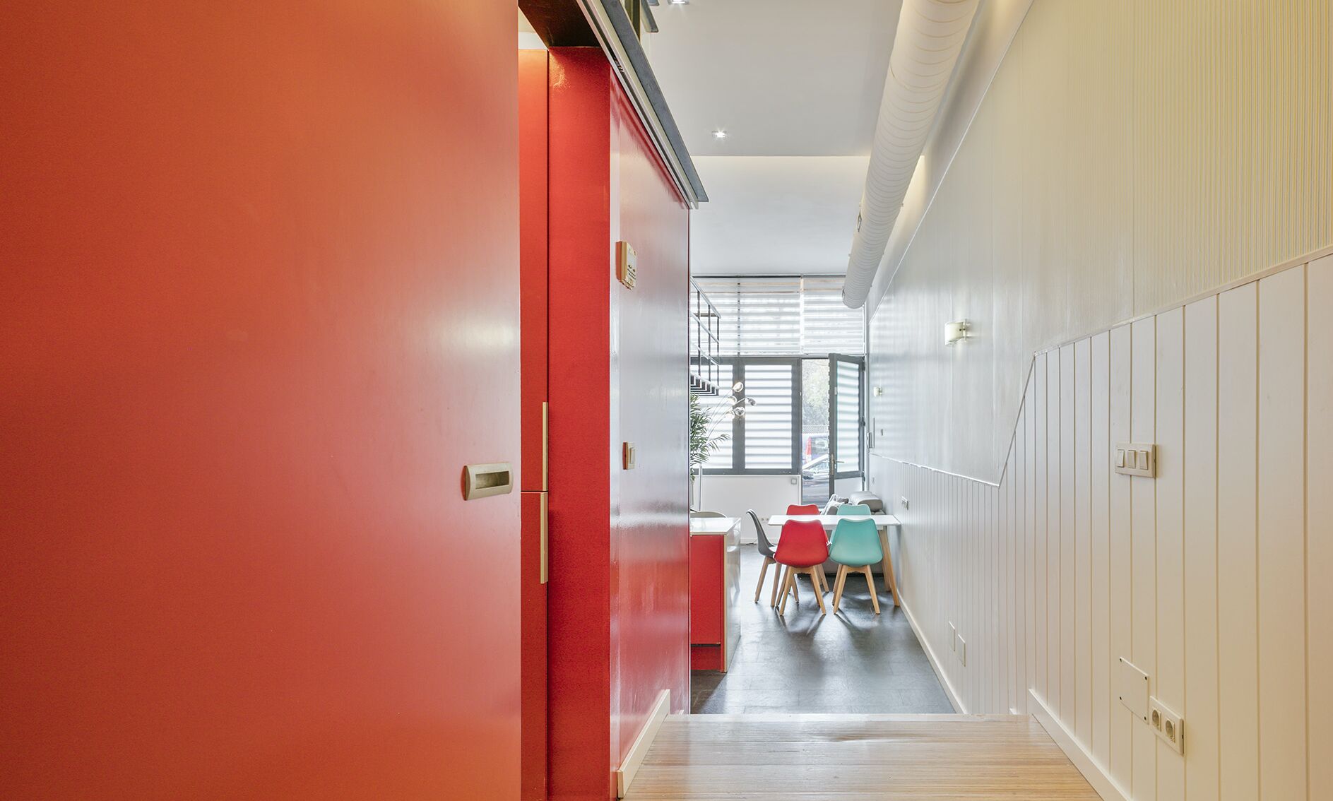 A hallway with a red door leading to a dining area with a table and chairs.
