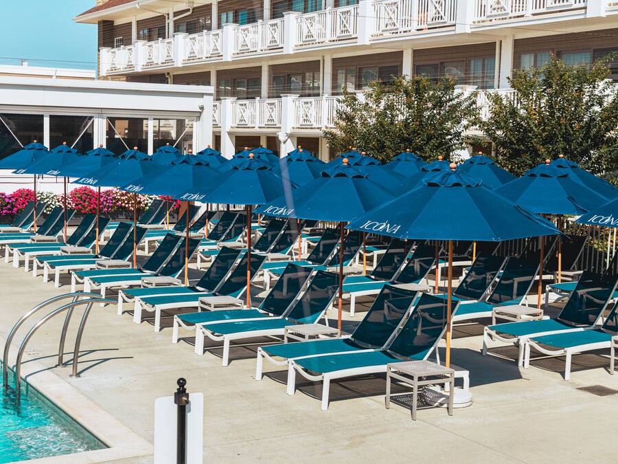 A poolside area with rows of sun loungers and blue umbrellas, a swimming pool, and a building with balconies.