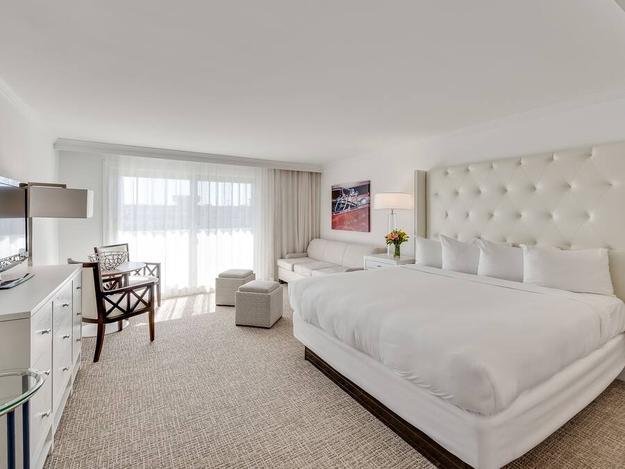 A modern hotel room with a large bed, a flat-screen TV, a desk with a chair, and a window with sheer curtains.