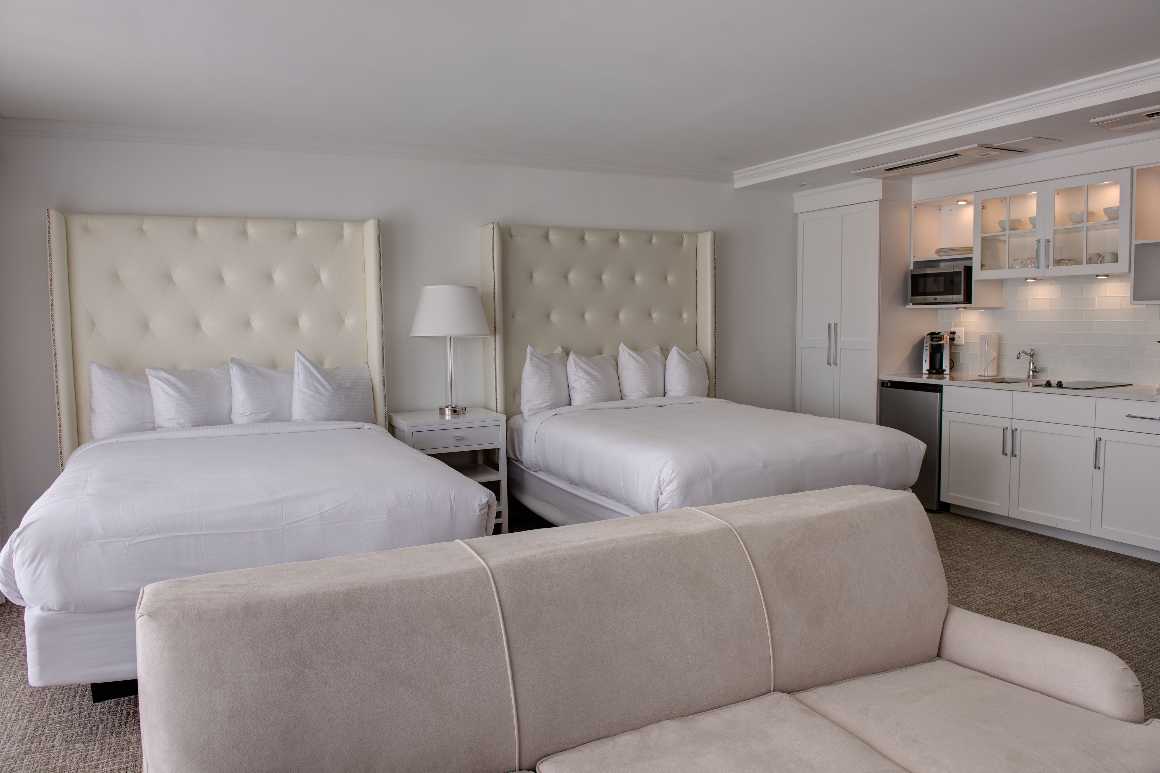 A hotel room with two beds, a sofa, a nightstand, a lamp, and a kitchenette.