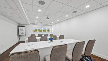 A conference room with a long table, six chairs, a projector, and a window with a view of trees.