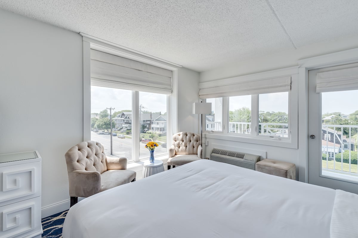 A hotel room with a large bed, two armchairs, a small table with a vase of flowers, and large windows offering a view of the street and buildings.