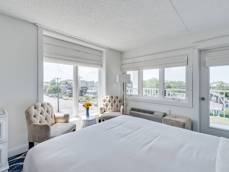 A hotel room with a large bed, two armchairs, a small table with a vase of flowers, and large windows offering a view of the street and buildings.