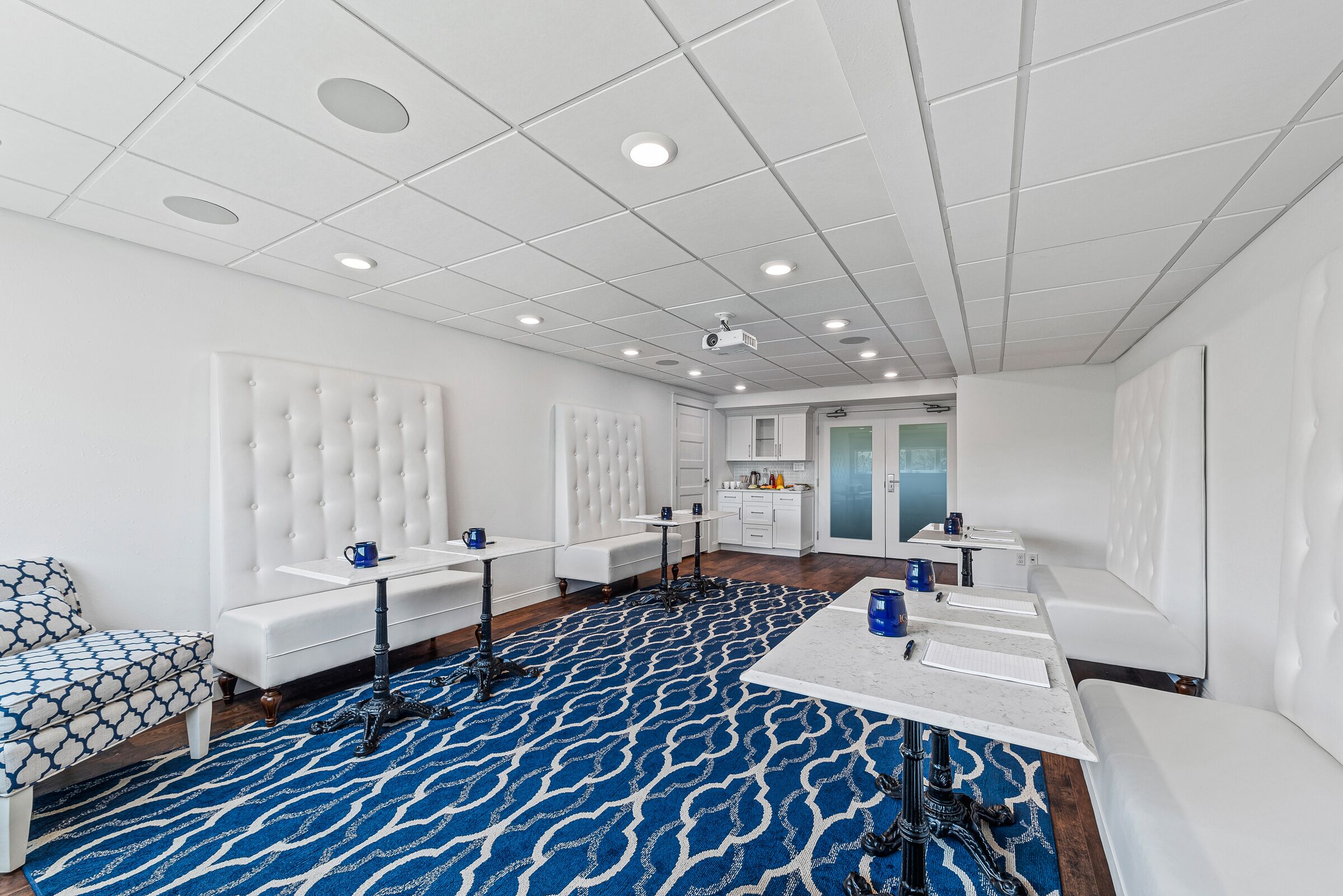 A modern hotel room with a large bed, a seating area with a patterned ottoman, a dining table, and a kitchen area.
