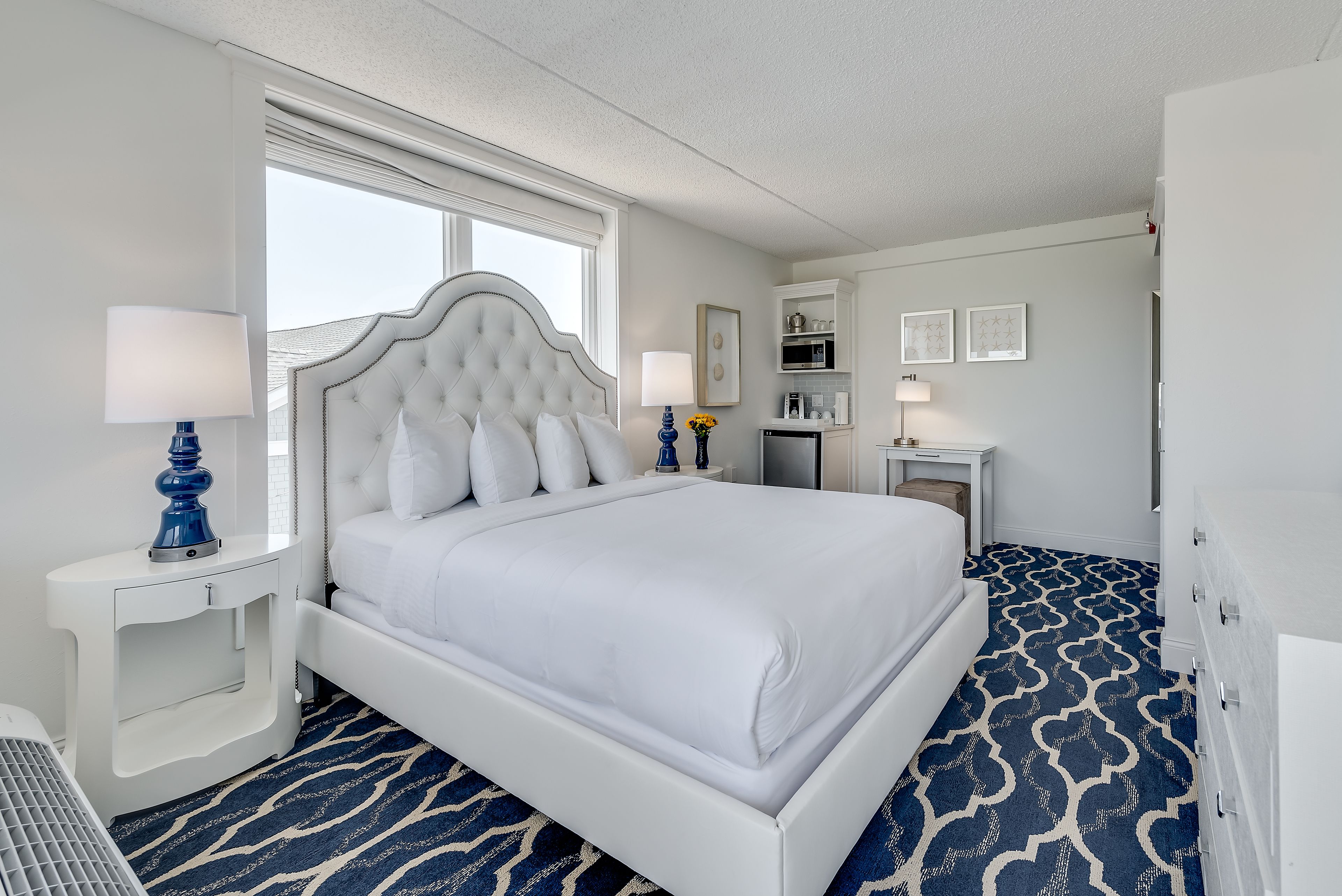 Une chambre d’hôtel avec un grand lit, des murs blancs, une fenêtre, une petite kitchenette et un tapis à motifs.
