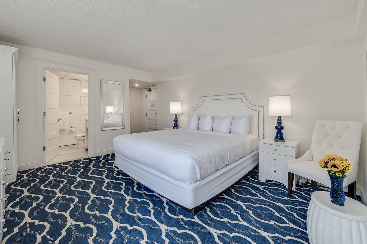 A hotel room with a large bed, a nightstand with a lamp, a chair with a vase of flowers, and a bathroom visible through an open door.