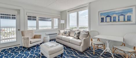 A spacious room with a large window overlooking the beach, a white sofa, a small table, and a patterned rug.