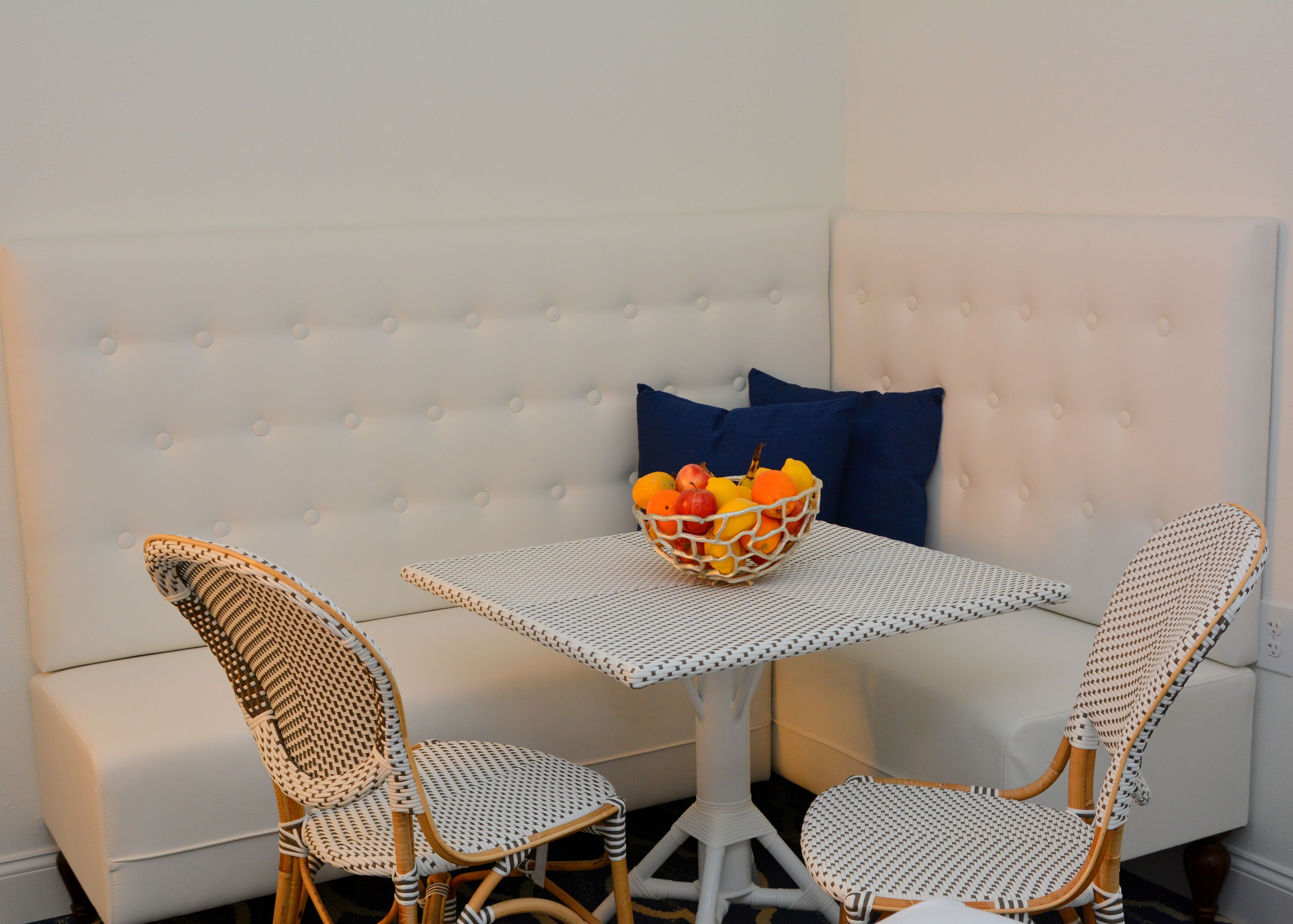 A corner seating area with a white cushioned sofa, a small white table, and two wicker chairs. A fruit bowl is placed on the table.