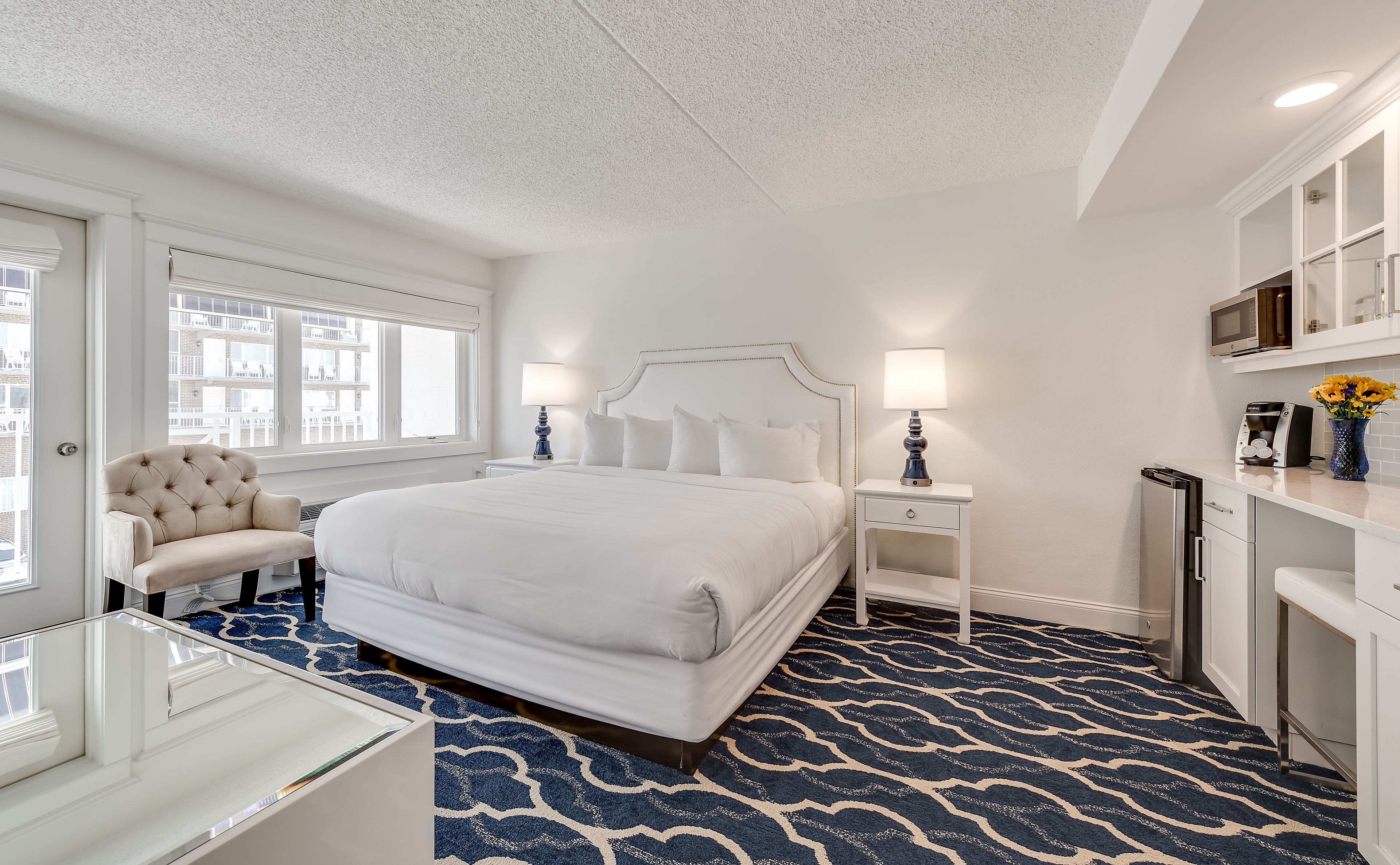 A hotel room with a large bed, a sitting chair, a desk, and a small kitchenette.